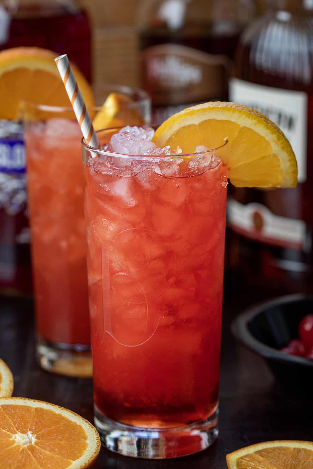 Alabama Slammer is an alcoholic beverage made with Southern Comfort, sloe gin, amaretto, and orange juice, shaken and poured over a glass of crushed ice. Top off the drink with orange slices and maraschino cherries for garnish. It is a refreshing cocktail that can also be served as a shot! Alabama Slammer Cocktail on a Cutting Board with Oranges and a Straw.