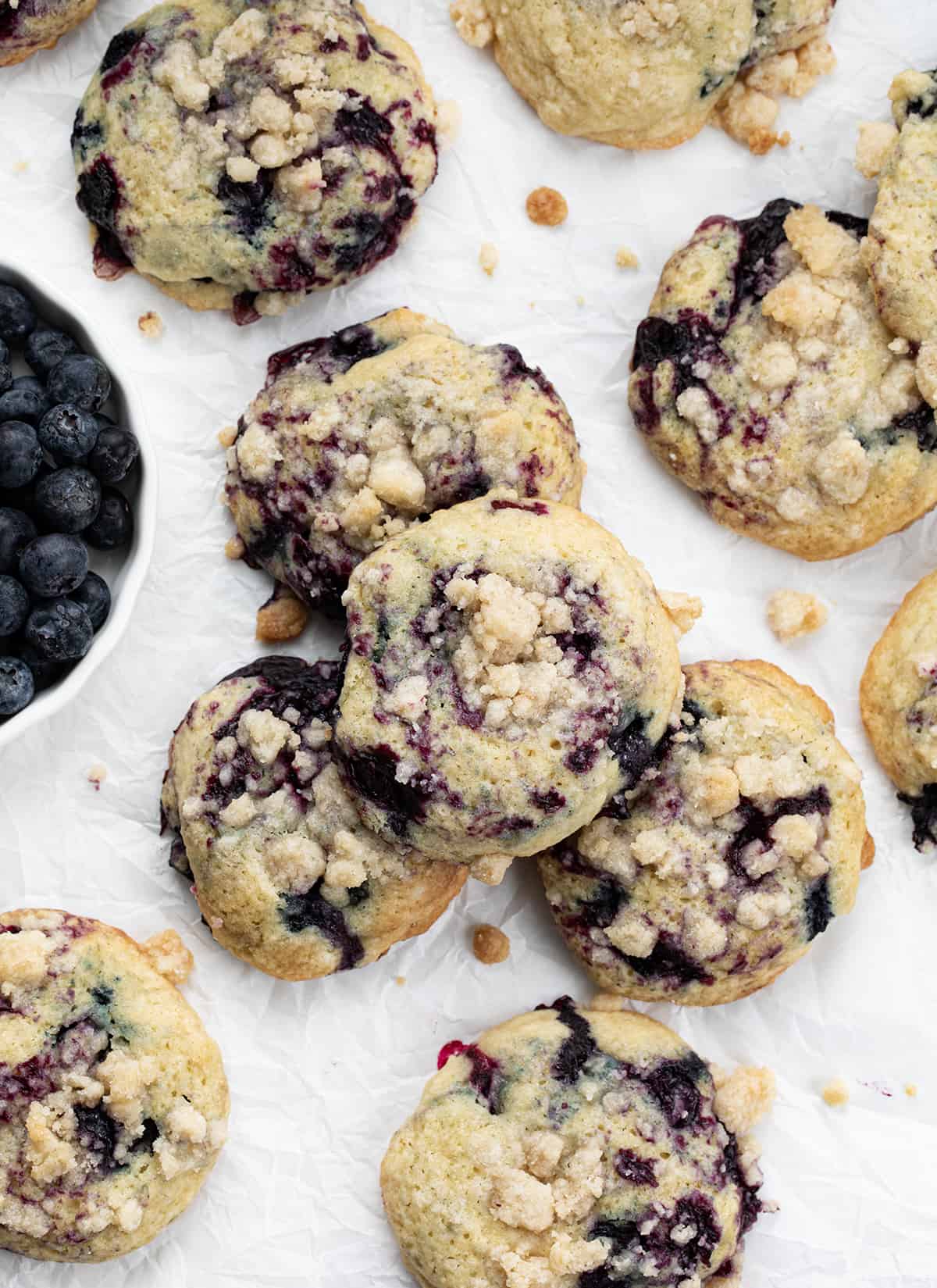 Blueberry Crumble Cake Cookies are soft, cake-like cookies with blueberry filling swirled inside and topped with a crumble topping. They are a delightful treat that combines the tenderness of a muffin or cake with the irresistible texture of a crumbly topping. They are truly a heavenly delight for blueberry lovers. Blueberry Crumble Cake Cookies on White Parchment Next to Fresh Blueberries.
