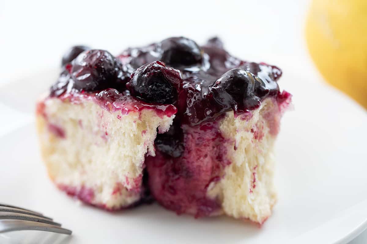 Blueberry Lemon Skillet Rolls are super soft rolls baked with lemon curd and fresh blueberries, then topped with a homemade blueberry sauce. They are the perfect treat for breakfast, brunch, or anytime you are craving one! I used an overnight dough to make things really easy for busy people, but no fear, there are also instructions for quicker use One Blueberry Lemon Skillet Roll on a White Plate with a Lemon in the Background.