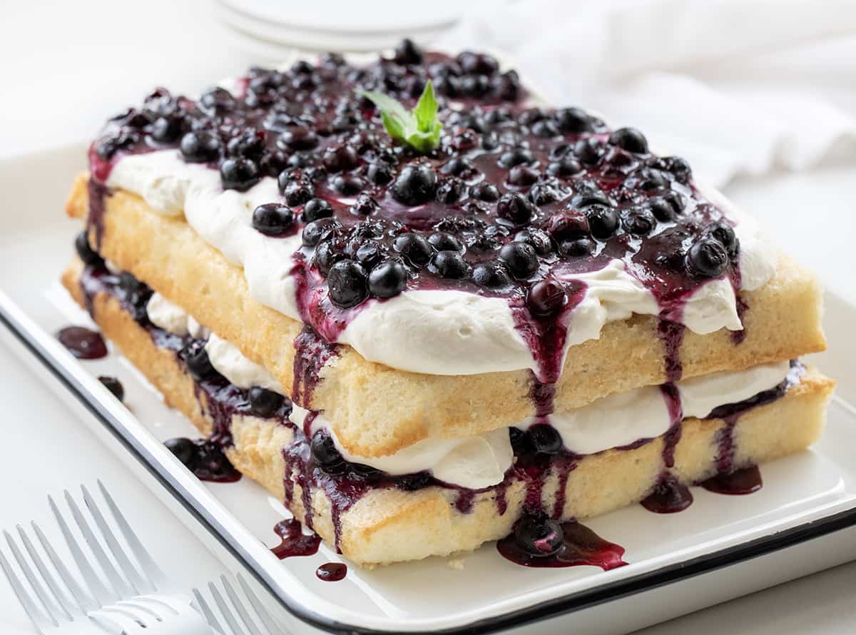 Sheet Pan Blueberry Shortcake Cake starts with a delicious white cake that is cut in half and layered with a homemade blueberry mixture and whipped cream cheese topping. It's a delightful dessert, combining the tender cake base with a sweet and tangy blueberry topping. The creamy whipped topping adds a luxurious and creamy element to the dessert, making it a perfect treat for any occasion. Whole Sheet Pan Blueberry Shortcake Cake on a White Counter.