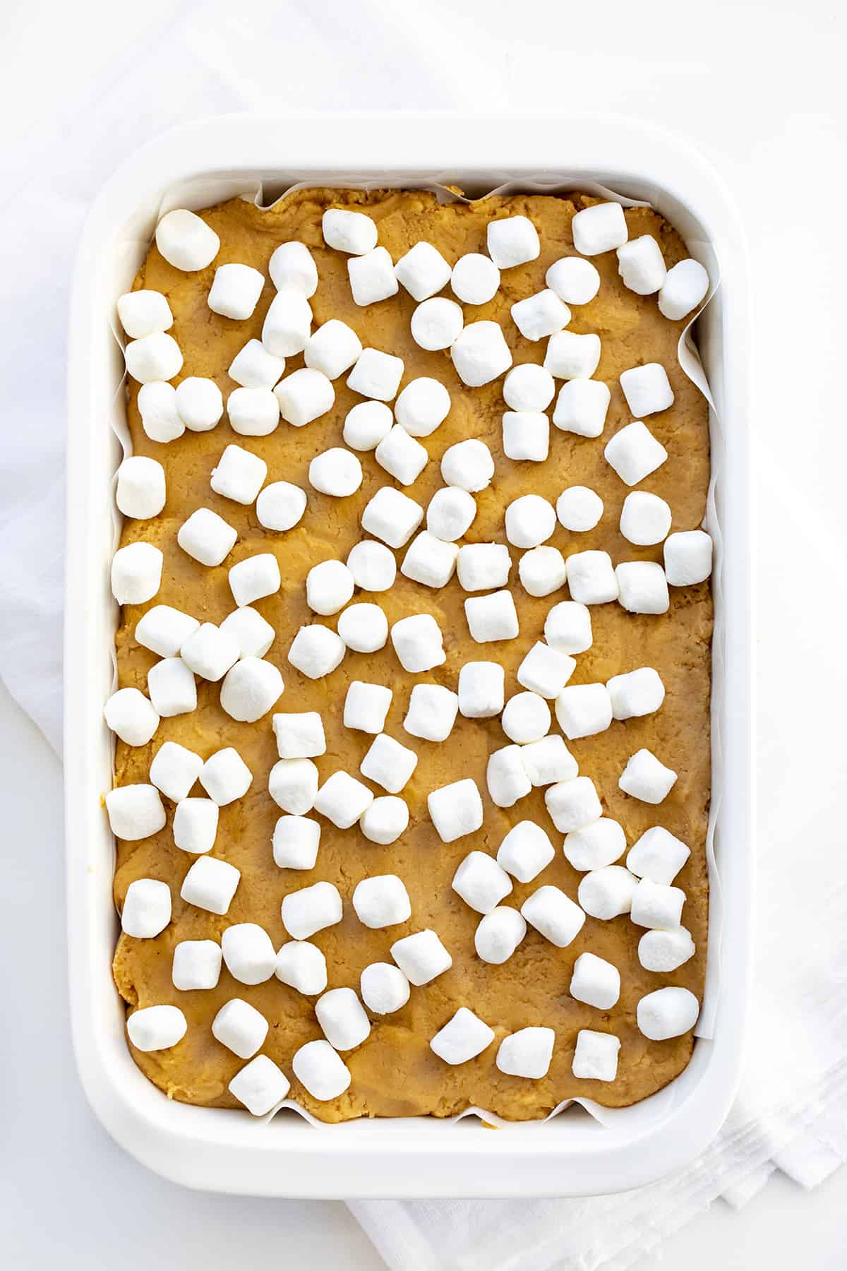 This fudge was inspired by a fluffernutter sandwich, which is a sandwich made with peanut butter and marshmallow fluff. Fluffernutter Fudge is two layers of melted peanut butter and white chocolate with a layer of marshmallow fluff in between. If you love peanut butter, do not miss out on this recipe! Pan of Completed Fluffernutter Fudge that Shows Peanut Butter Fudge Topped with Mini Marshmallows on White Counter.