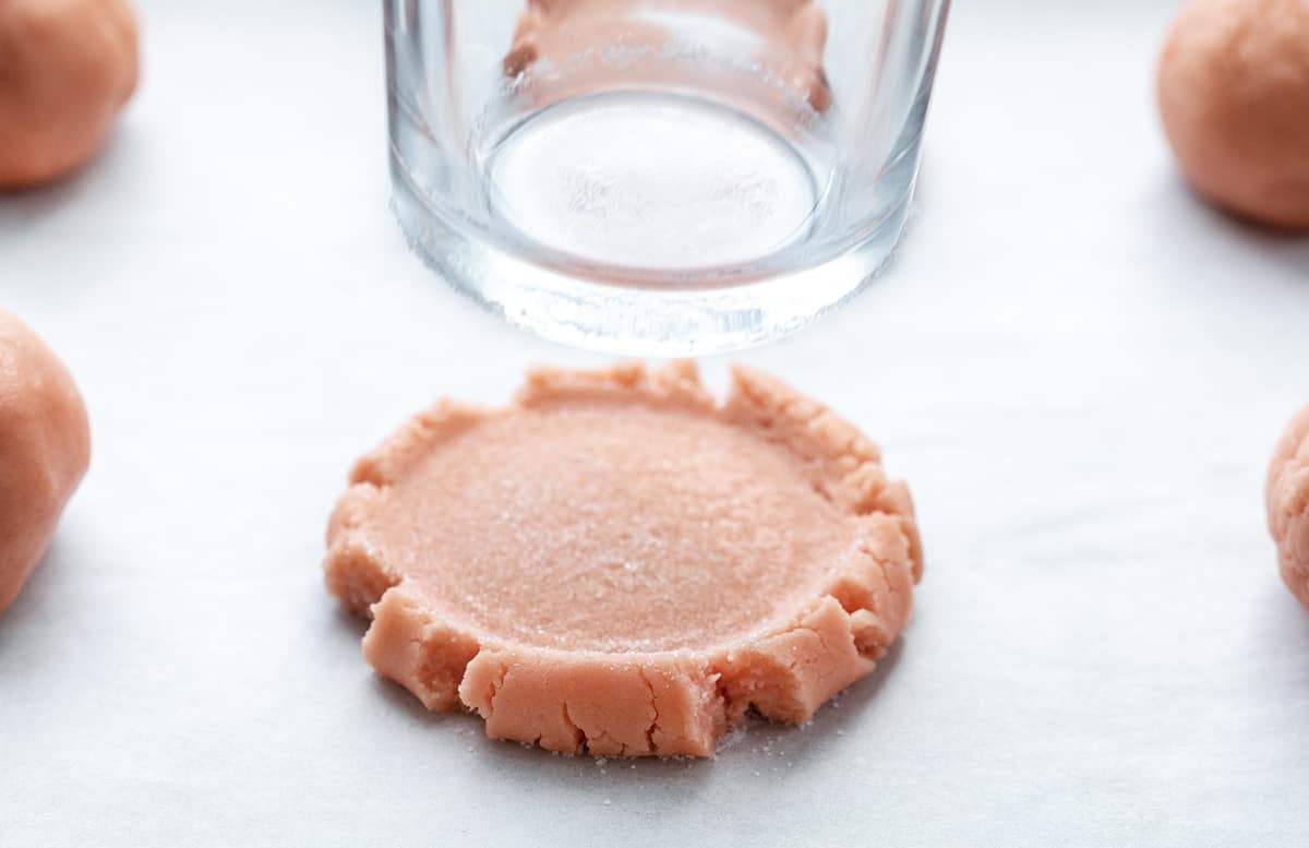Pink Velvet Sugar Cookies with Vanilla Meringue are soft sugar cookies tinted to a light pink color and frosted with an easy, velvety Swiss Meringue buttercream. Pressing a Glass into a Pink Velvet Sugar Cookie.
