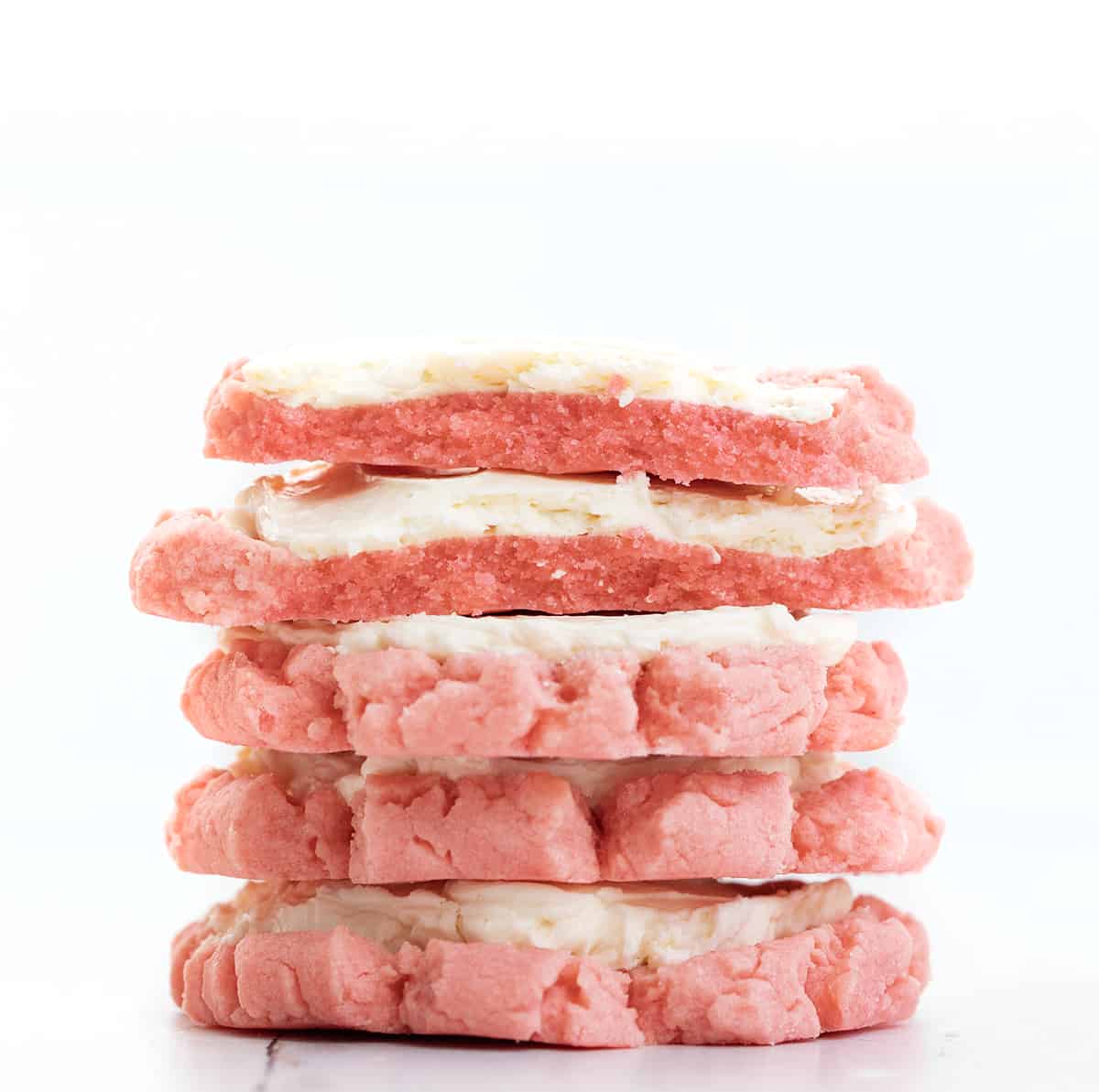 Pink Velvet Sugar Cookies with Vanilla Meringue are soft sugar cookies tinted to a light pink color and frosted with an easy, velvety Swiss Meringue buttercream. Stack of Pink Velvet Sugar Cookies with Easy Swiss Meringue Frosting with the Top Cookies Broken in Half.