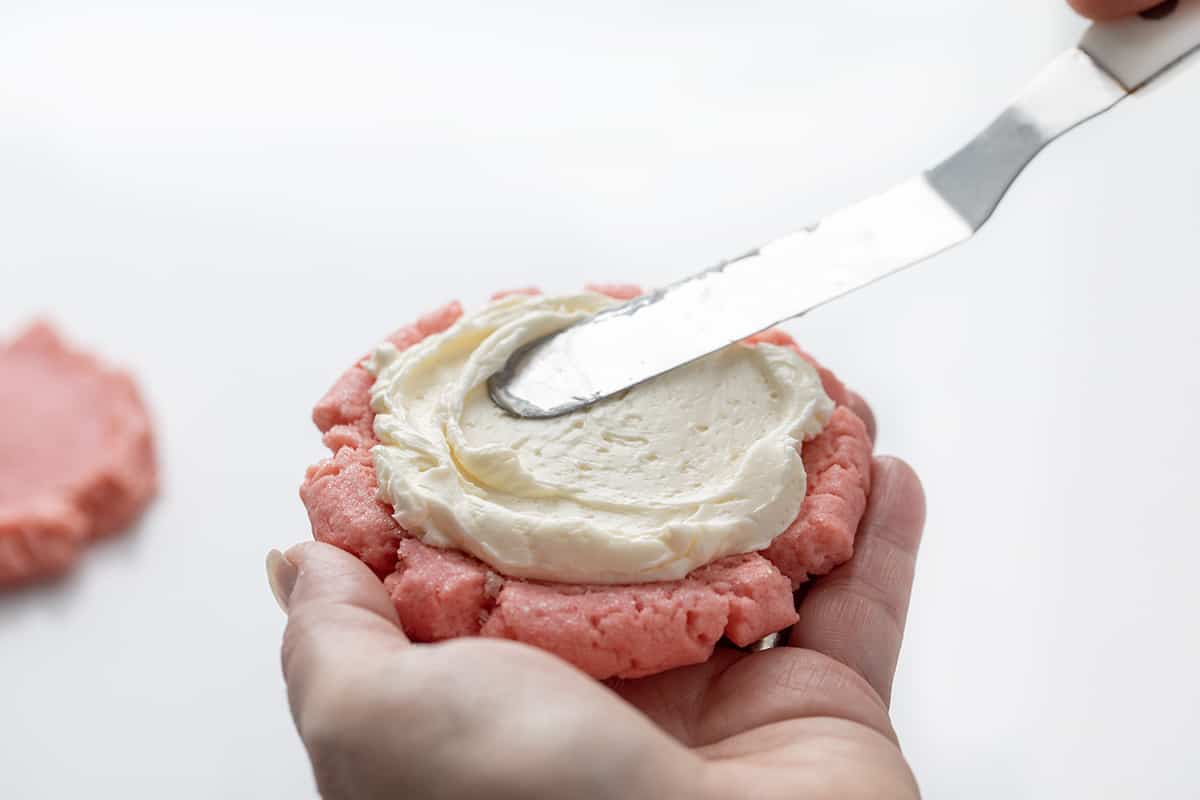 Pink Velvet Sugar Cookies with Vanilla Meringue are soft sugar cookies tinted to a light pink color and frosted with an easy, velvety Swiss Meringue buttercream. Smoothing Swiss Meringue Frosting Over a Pink Velvet Cookie.