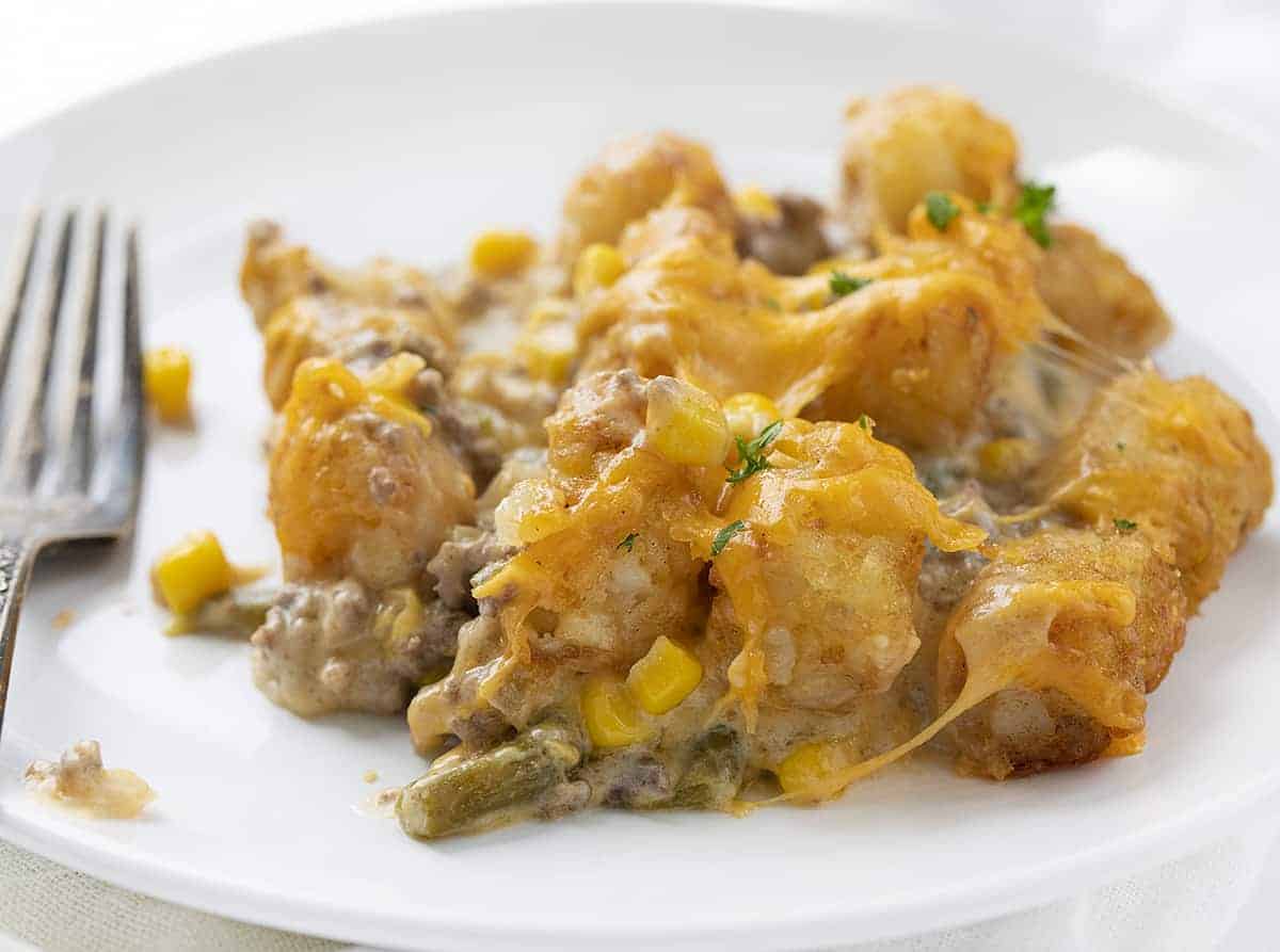 Tater Tot Hotdish is a quick and easy comfort food staple recipe all over the Midwest! This hotdish is full of meat, veggies, soup, cheese, and topped with tater tots for a super satisfying meal! Tater Tot Hotdish, Tater Tot Casserole, How to Make Hotdish, Casserole, Tater Tot Recipes, Dinner, Supper, Recipes, Dinner Ideas, Midwest Recipes, i am baker, iambaker White Plate with Tater Tot Hotdish on it and a Fork Next to it